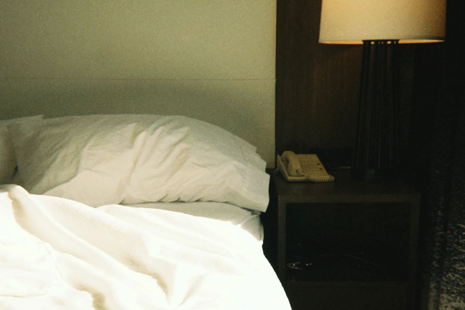 An unmade hotel bed with a rumpled pillow, a small lit lamp glowing on the nightstand beside it in a dim room