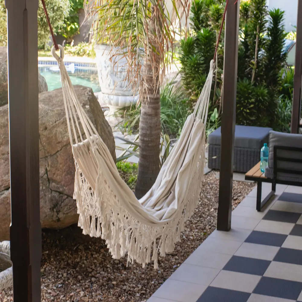 A cream fringed hammock on a covered veranda beside a garden with a palm, large boulders and outdoor seating