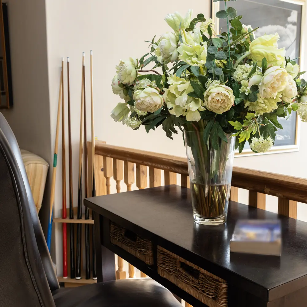 A vase of white roses and peonies on a dark hall table beside a wooden banister and a rack of cues