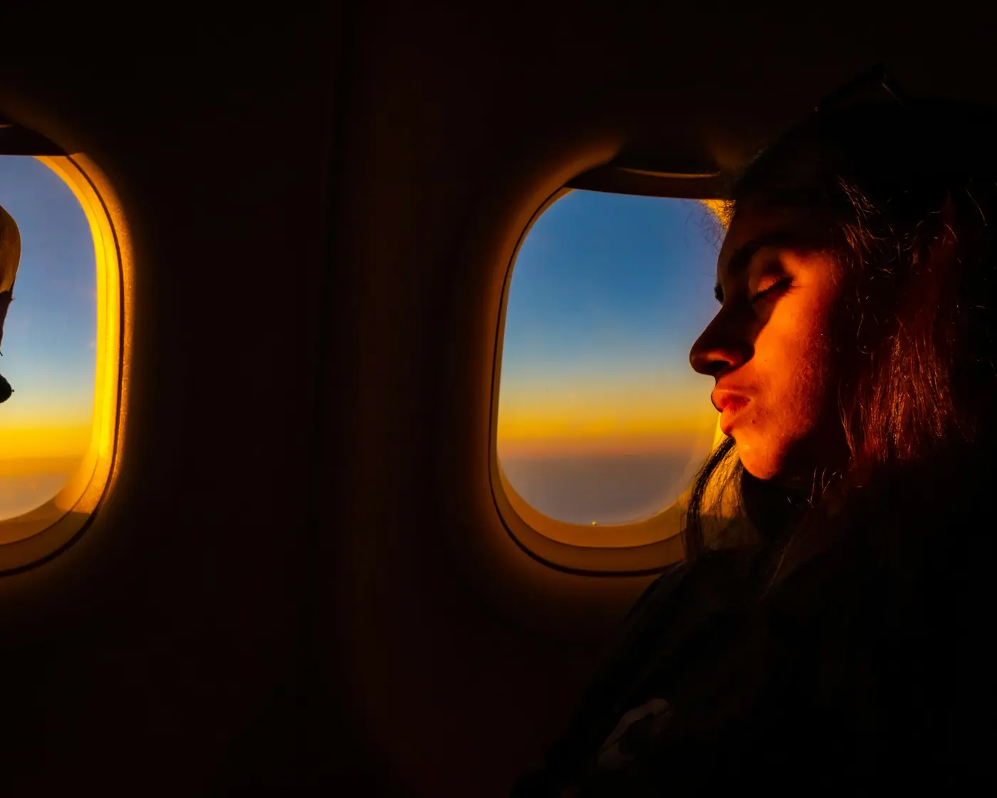 A woman in a window seat looks out at a golden sunset above the clouds on a long flight