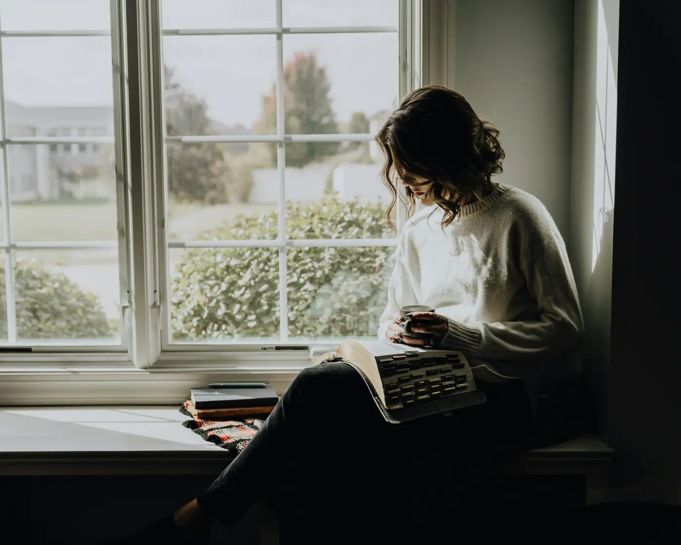 A woman in a cream jumper sits in a window seat with a mug in her hand and a typewriter on her lap