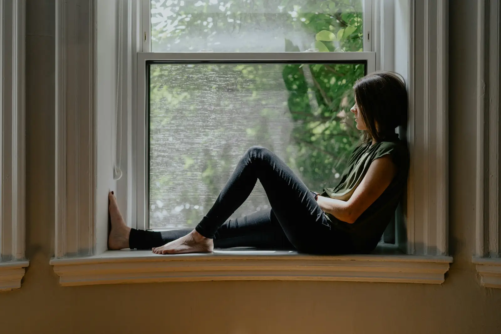 A woman sitting barefoot on a window sill, looking out at green trees
