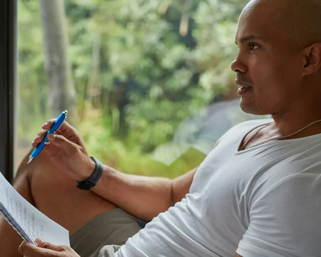 Man in a white t-shirt holding a pen and reading a paper form by a bright window
