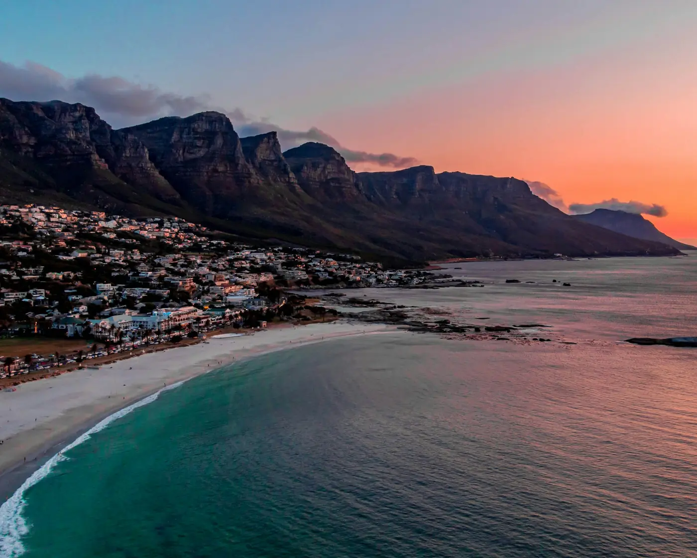 A white-sand bay curving below a long mountain ridge in Cape Town at sunset, pink and orange sky over the sea