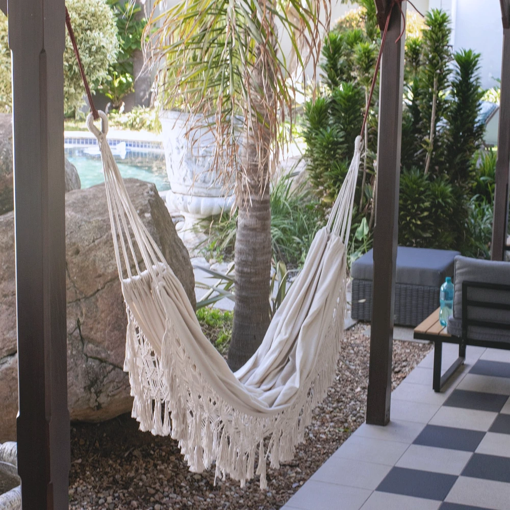 An empty cream macrame hammock strung between a palm tree and a boulder in a sunlit garden