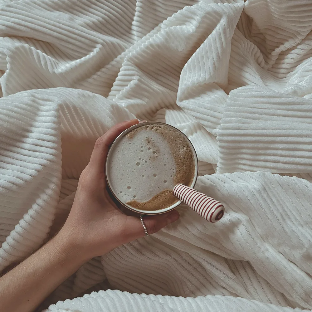 A hand holding a mug of coffee over a cream corduroy blanket