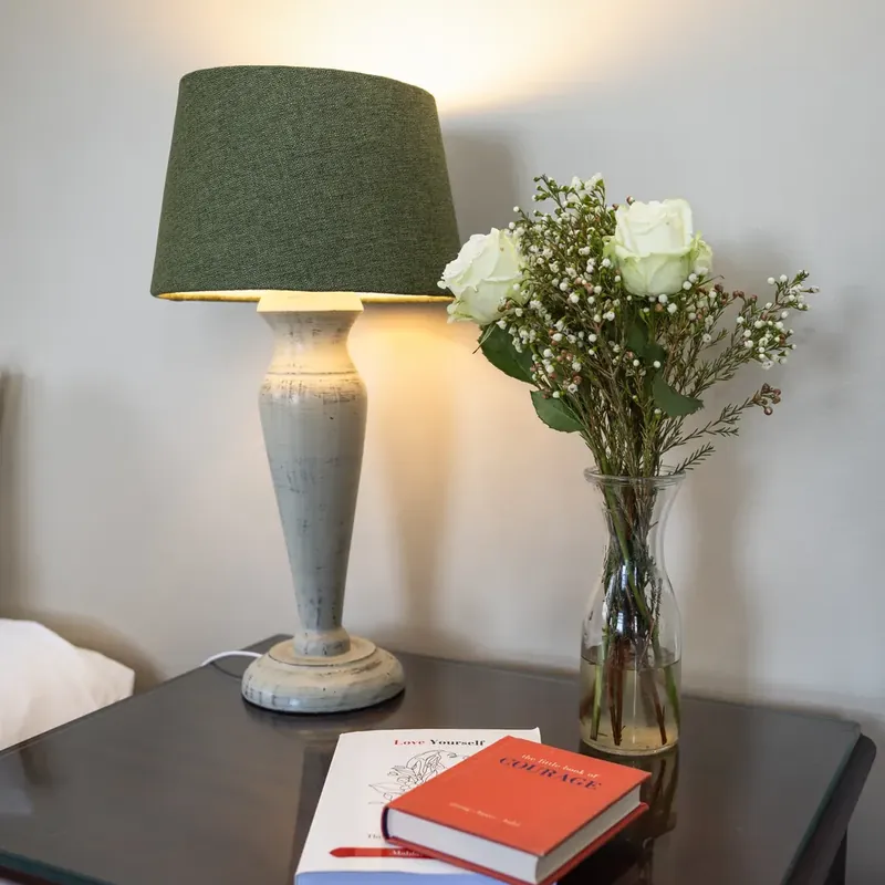 A table lamp with a green shade beside a vase of white roses and a red book on a dark bedside table