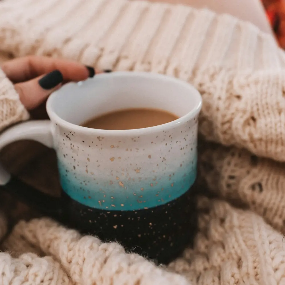 Hands around a speckled turquoise mug of milky tea, wrapped in a cream knitted blanket