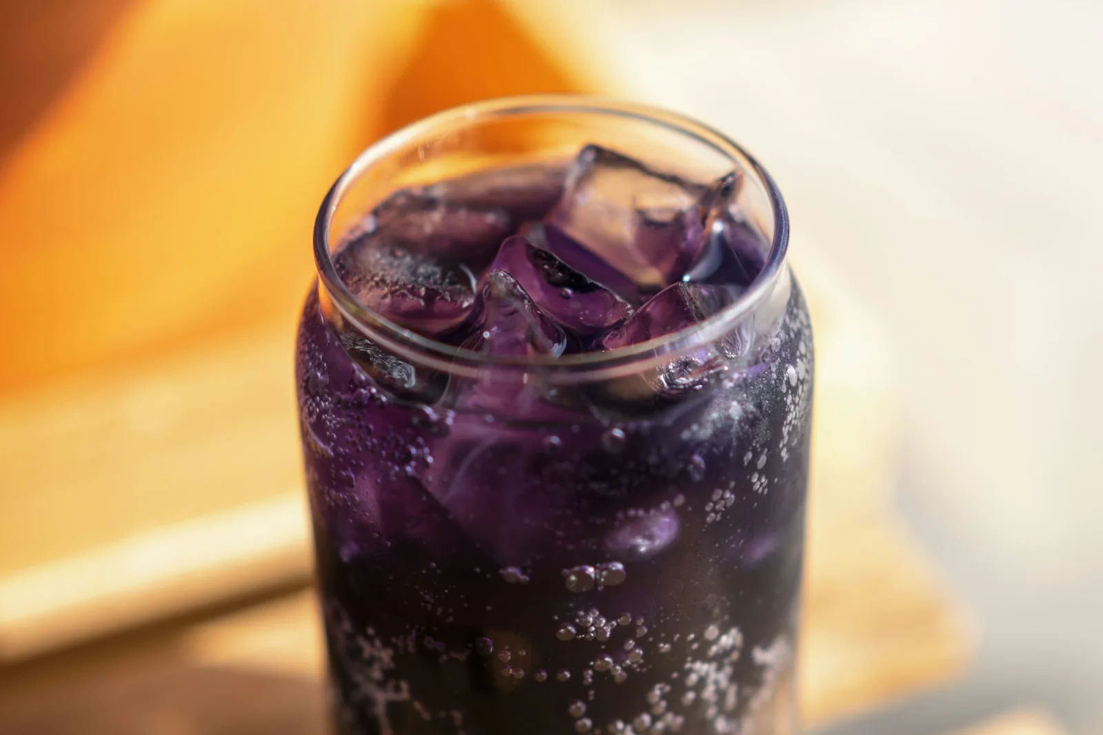 A glass of fizzy dark purple drink with ice cubes on a wooden board in warm light