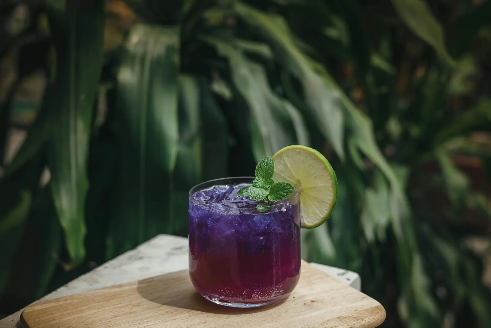 A glass of purple drink with ice, a sprig of mint and a slice of lime on a wooden board among green leaves