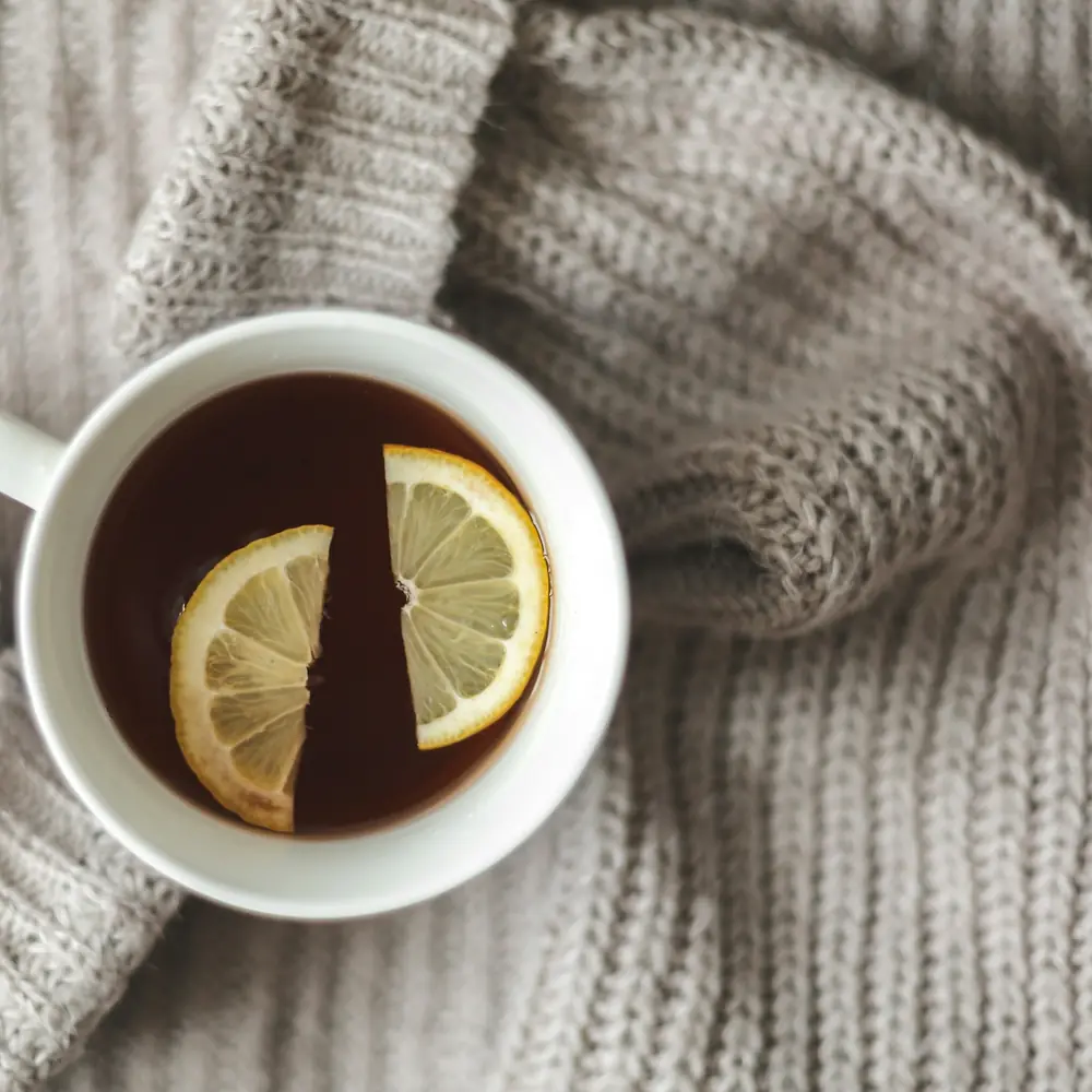 A white cup of black tea with lemon slices on a knitted grey sweater, seen from above