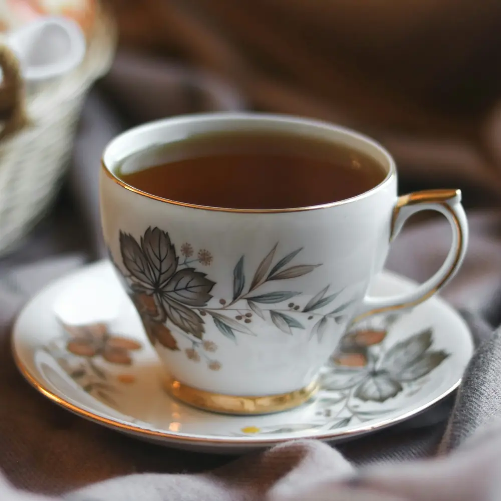 A white china cup of tea with a gold rim on a soft blanket beside a wicker basket
