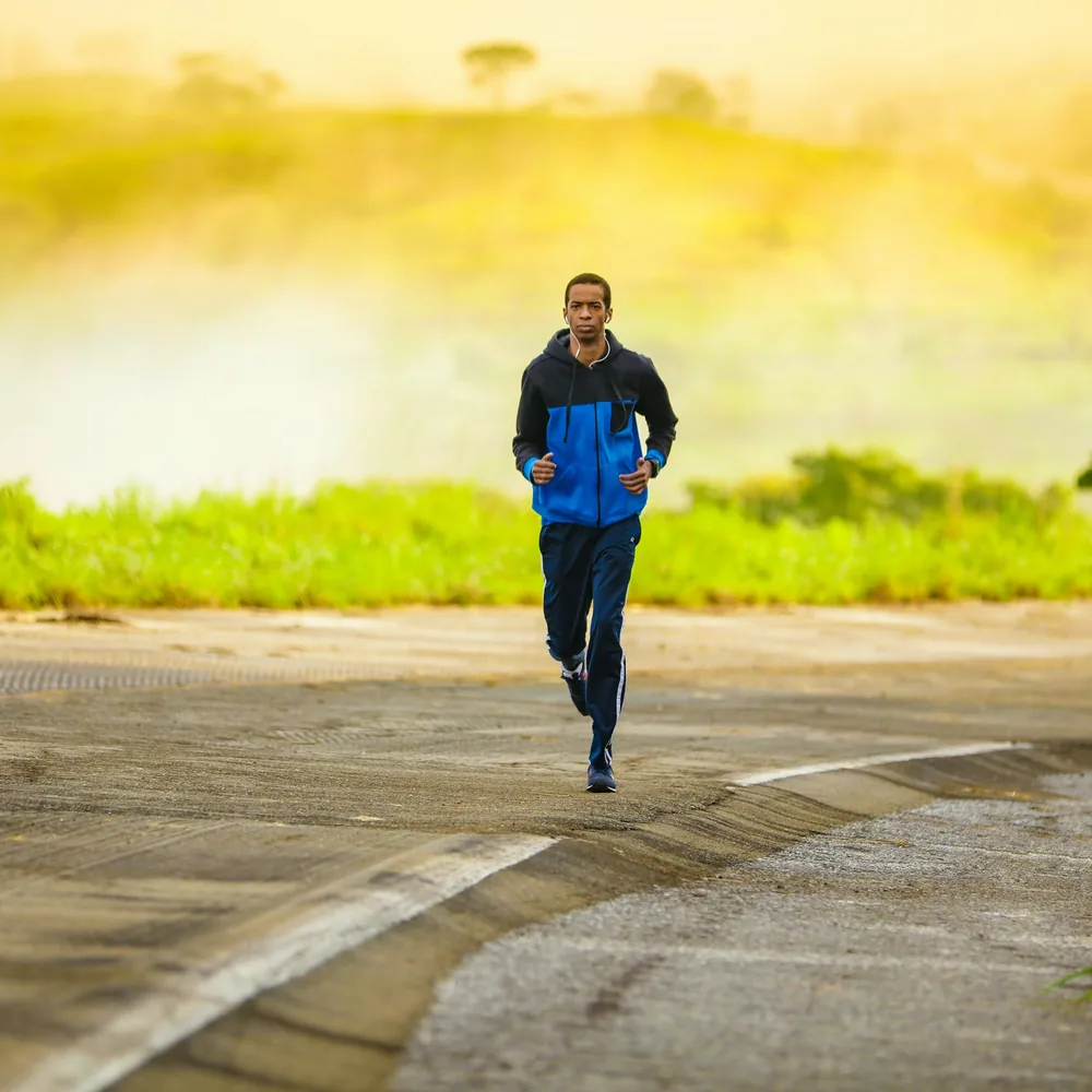 Man in a blue jacket jogging along a road through green fields in warm light